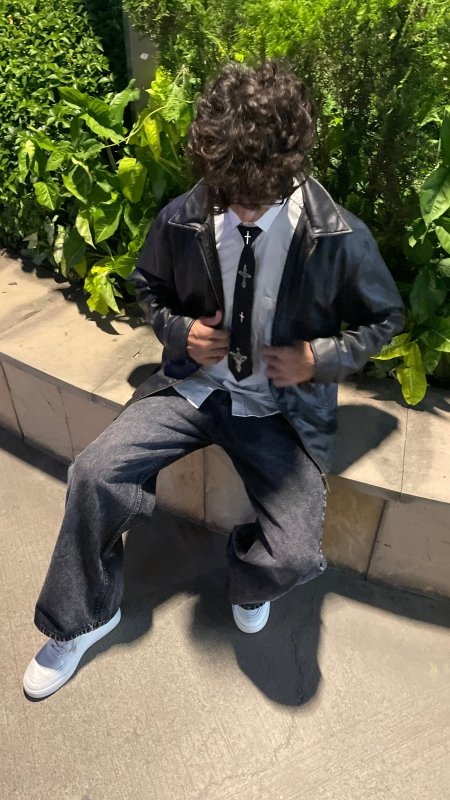 The Black Vow Tie - StreetgenA person sitting outdoors on a stone ledge wearing a black leather jacket, white shirt, black tie, dark wide-leg jeans, and white sneakers, with green plants in the background.